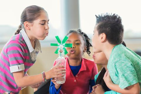 Four children blowing on small windmill placed inside plastic bottle.