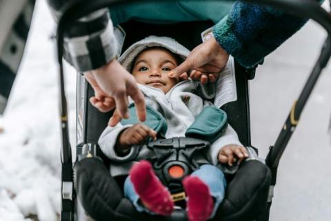 Baby with gray hoodie, blue pants and red socks smiling in stroller.