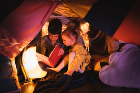 Two children reading in a lamp-lit blanket fort.