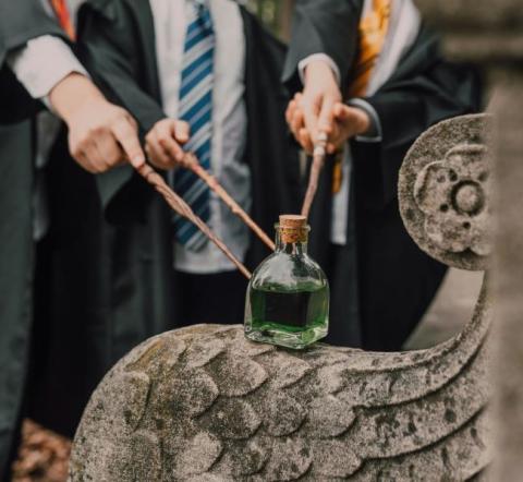 Hands of three boys dressed as wizards pointing their wands at a green potion.