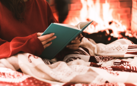 Woman reading under a blanket with lit fireplace in the background.