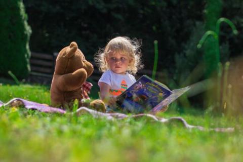 Child sitting with teddy bear on picnic blanket in a field, reading.