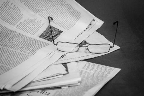 Newspaper pages spread along a surface from the center of the image to the left. A pair of glasses rests in the center on top of the pages.