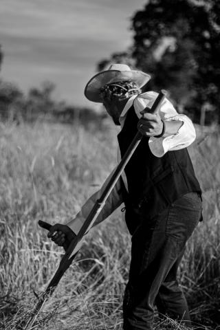 Older farmer with brimmed hat, vest and button down shirt, and jeans seen at an angle from behind. He is working in a field of wheat or grains with a two-handed sickle, reaping a harvest.