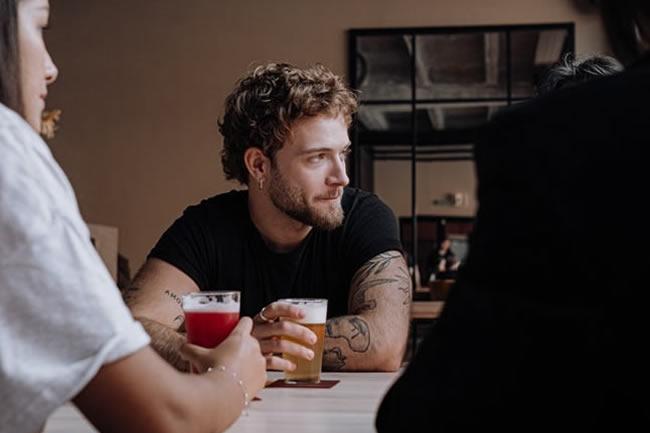 Adult male in black t-shirt sitting at a table with a glass of beer.