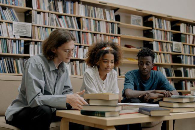 Three students perusing books on a table in a library.