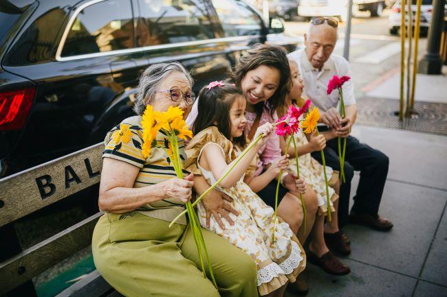 Grandparent, Parent, and Child sitting on bench in city holding flowers and laughing together.